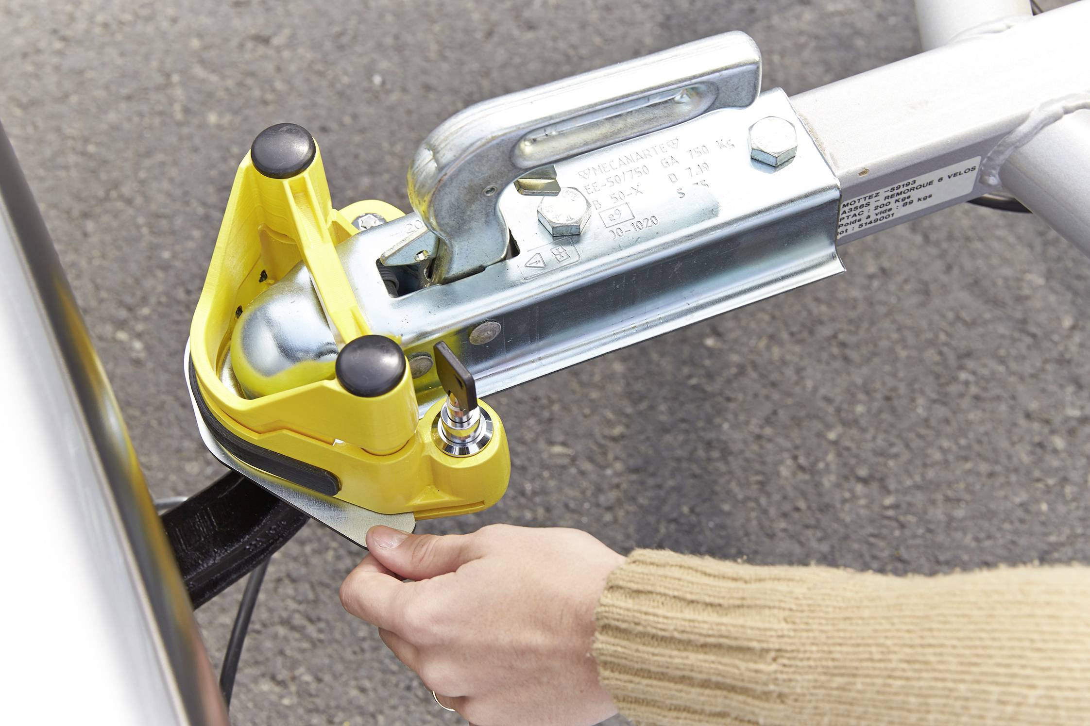A hand is attaching a yellow towbar to a trailer. Safety chain and locking mechanism are visible.