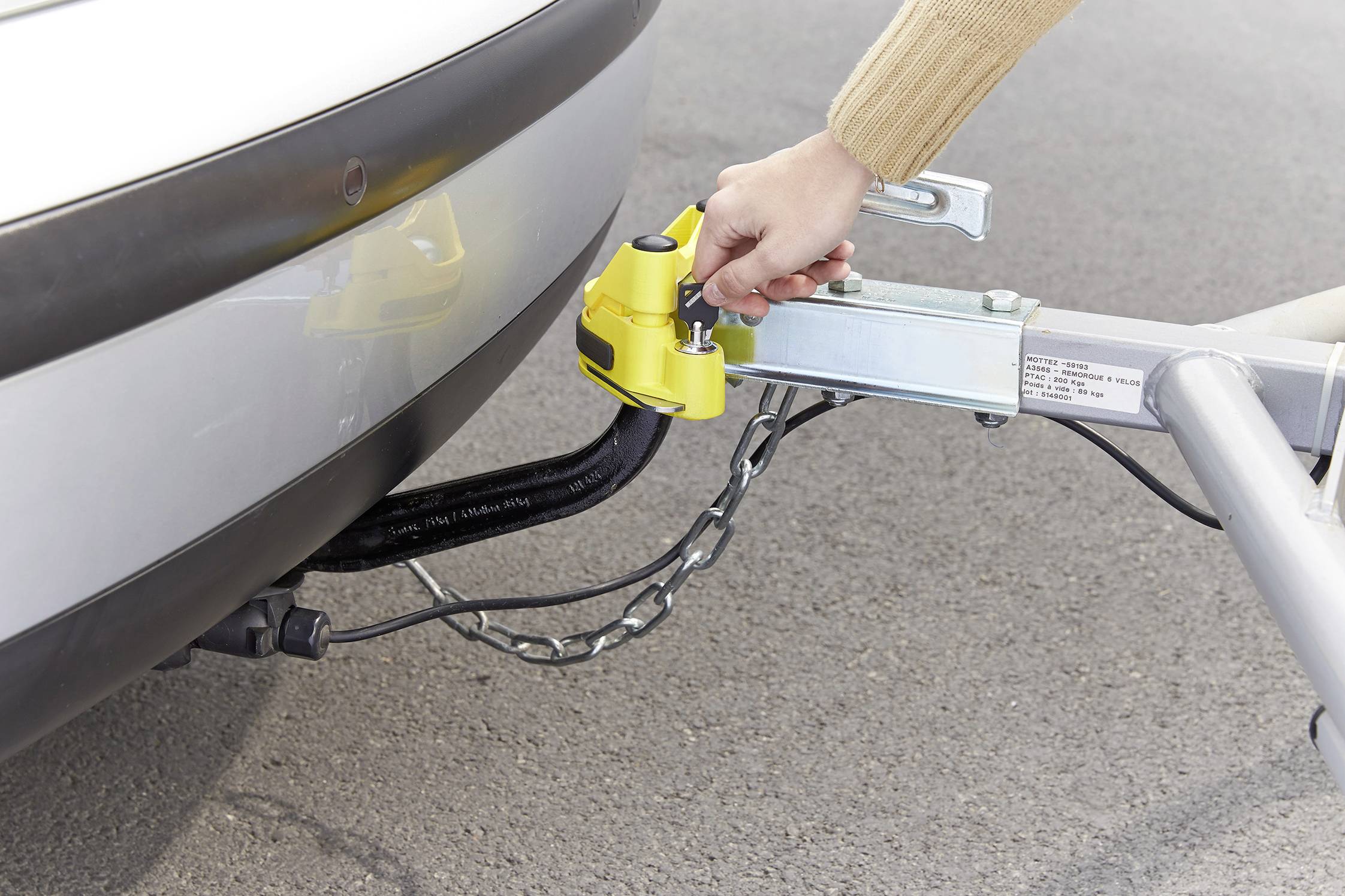 'A person secures a yellow coupling safety device to a vehicle's tow hook on an asphalted surface.'