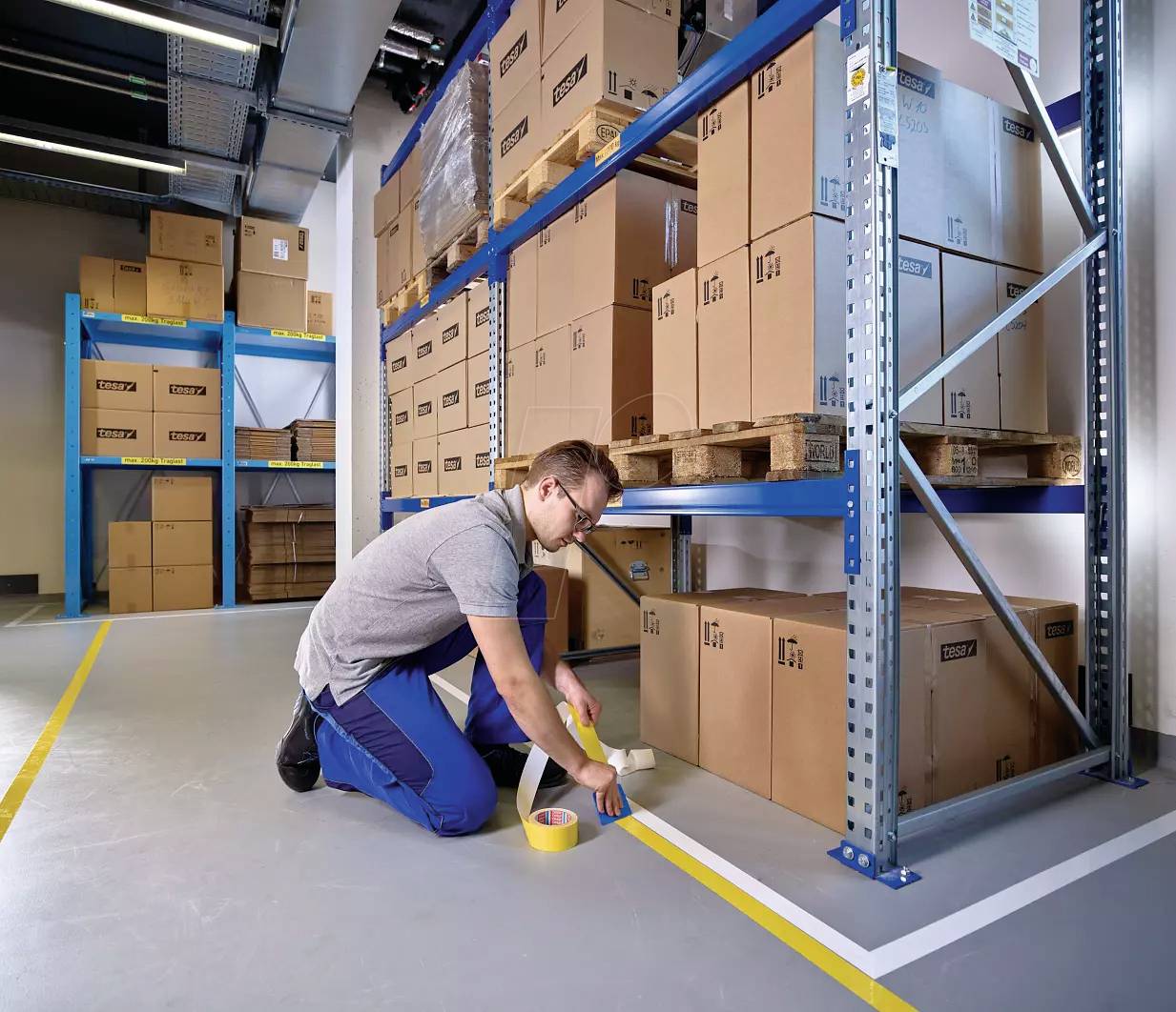 A person is kneeling in a warehouse and applying yellow adhesive tape to the floor to create markings. Storage shelves with cardboard boxes are visible in the background.