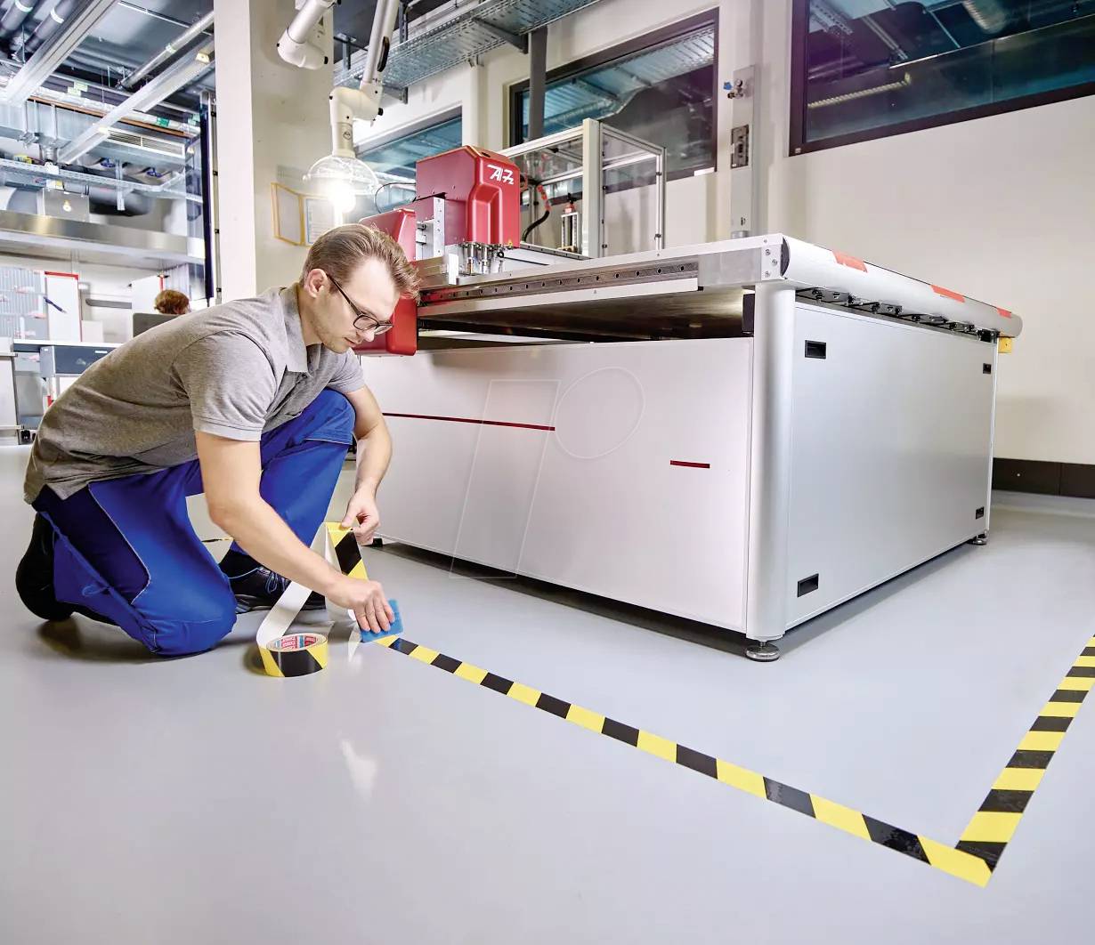 A person is applying black and yellow warning markings to the floor in an industrial space beside a large machine.