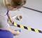 A person is sticking a black and yellow striped adhesive tape onto the floor. The tape indicates a safety marking.