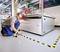 A man is marking the floor of a laboratory with black and yellow adhesive tape next to a large machine to create a safe working area.