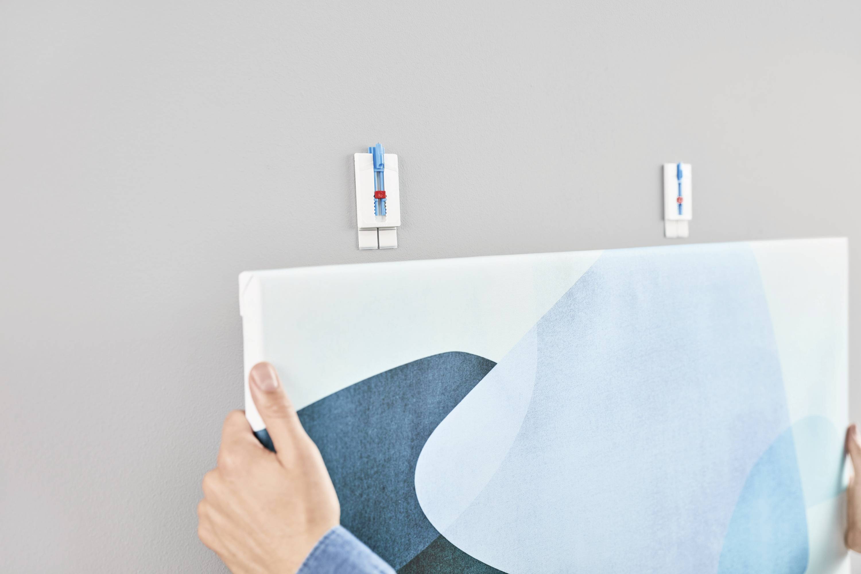 A person is hanging a picture on the wall. The picture is secured with two white mounting clips. A person's hands are visible.