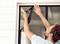 A man is installing a mosquito net on the window to keep insects out. He pulls the mesh taut and secures it carefully.