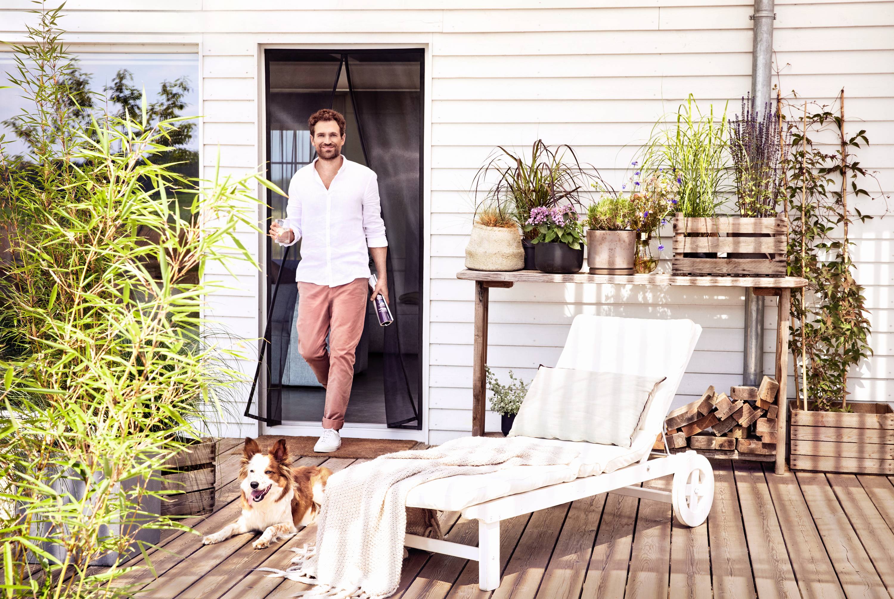 A man in casual clothing steps out onto a terrace from a house. A dog sits beside him. The terrace is decorated with plants and seating areas.