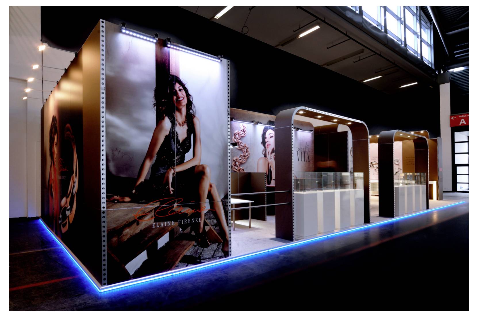 Fashion exhibition booth with large model posters, illuminated display counters, and blue LED floor lighting in a convention hall.