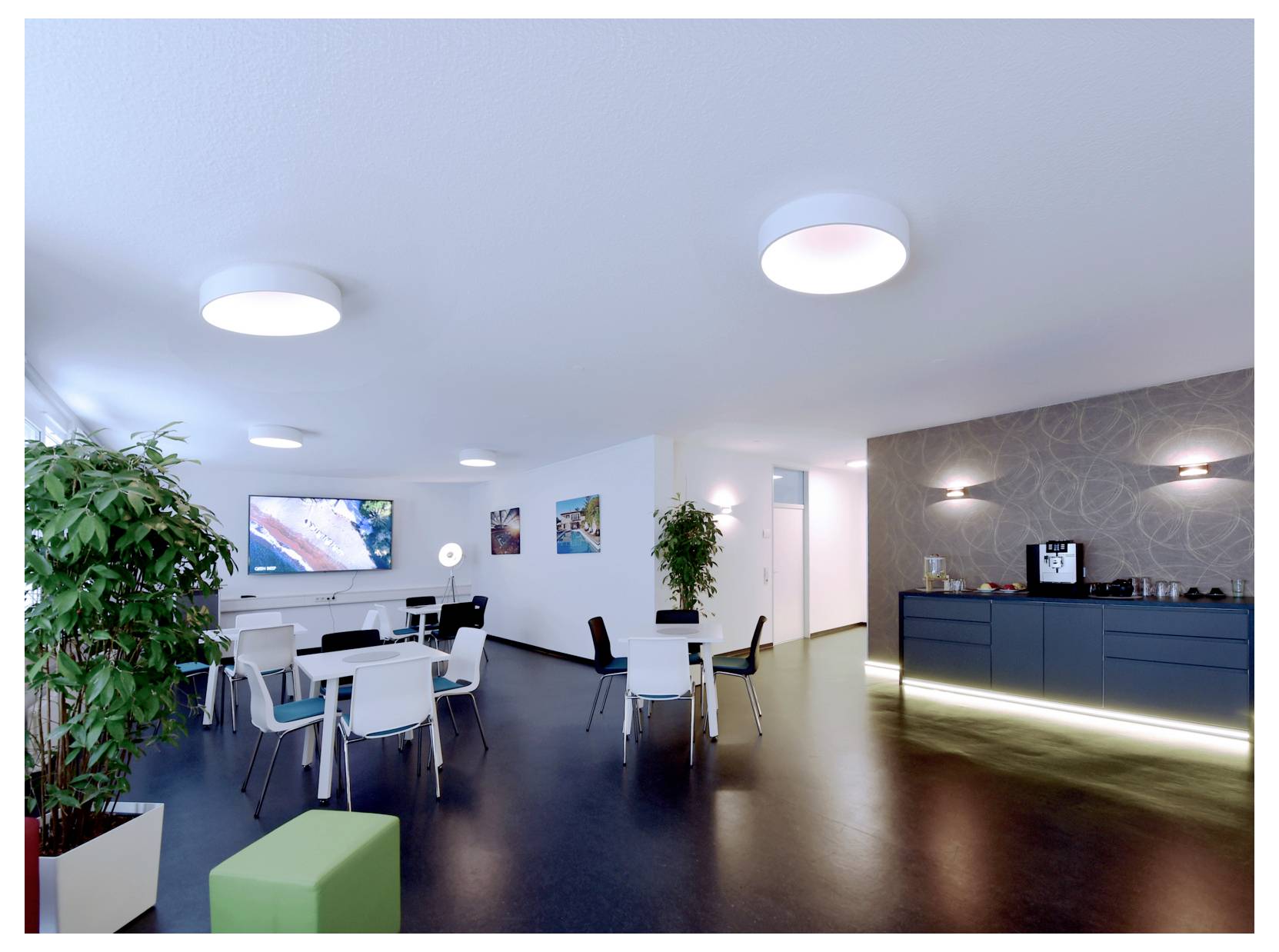 Modern office lounge with white tables and chairs, wall-mounted TV, potted plants, and a dark sideboard with lights along the right wall.