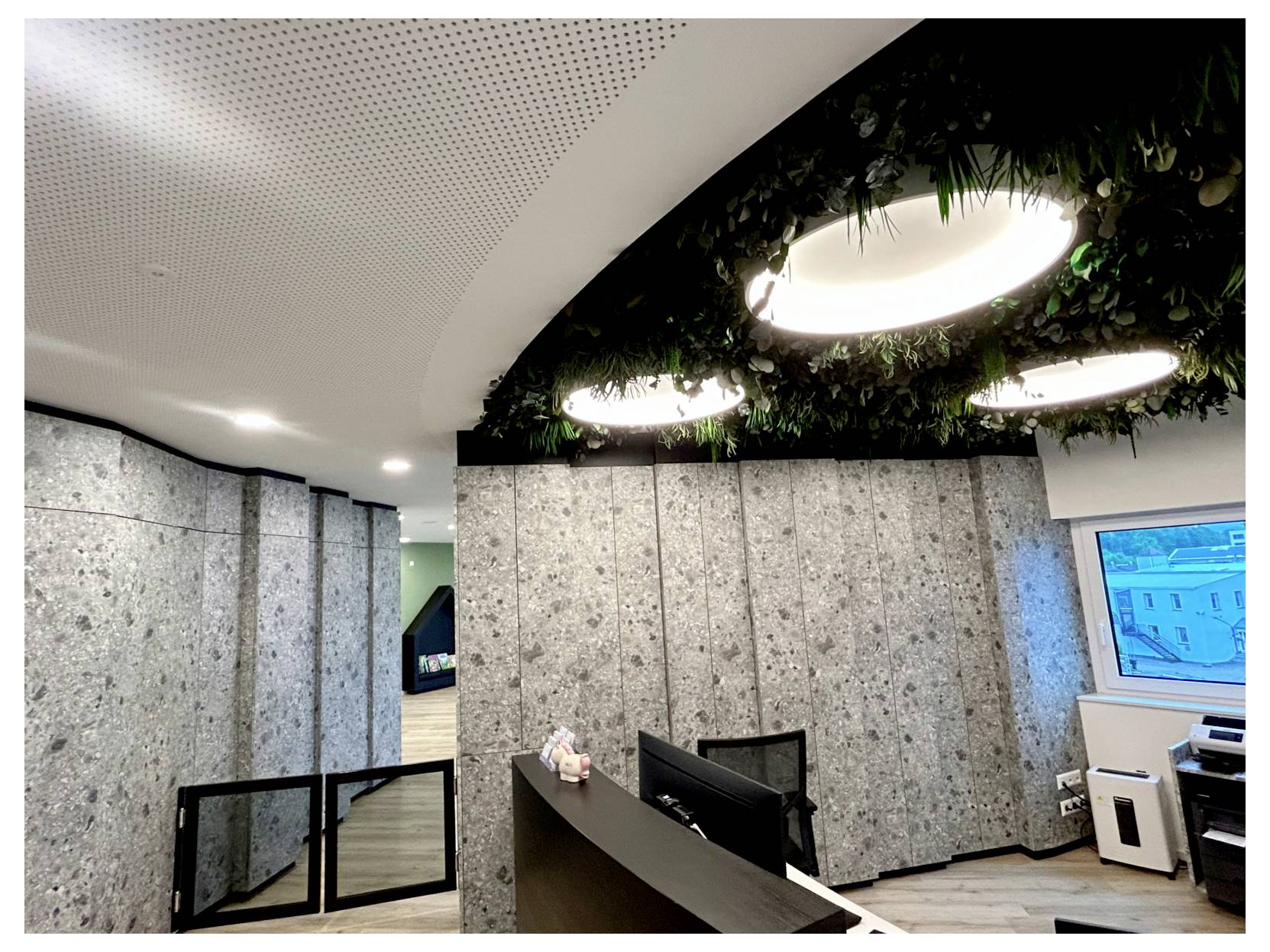 Modern office lobby/reception area with speckled stone walls, a curved ceiling with circular lights and greenery, and a reception desk with monitor.