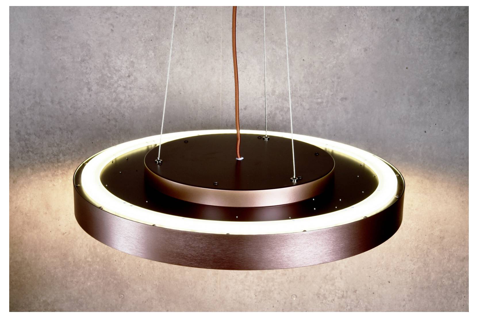 Modern circular pendant light with a glowing ring and dark central disk, suspended by thin cables against a plain wall.