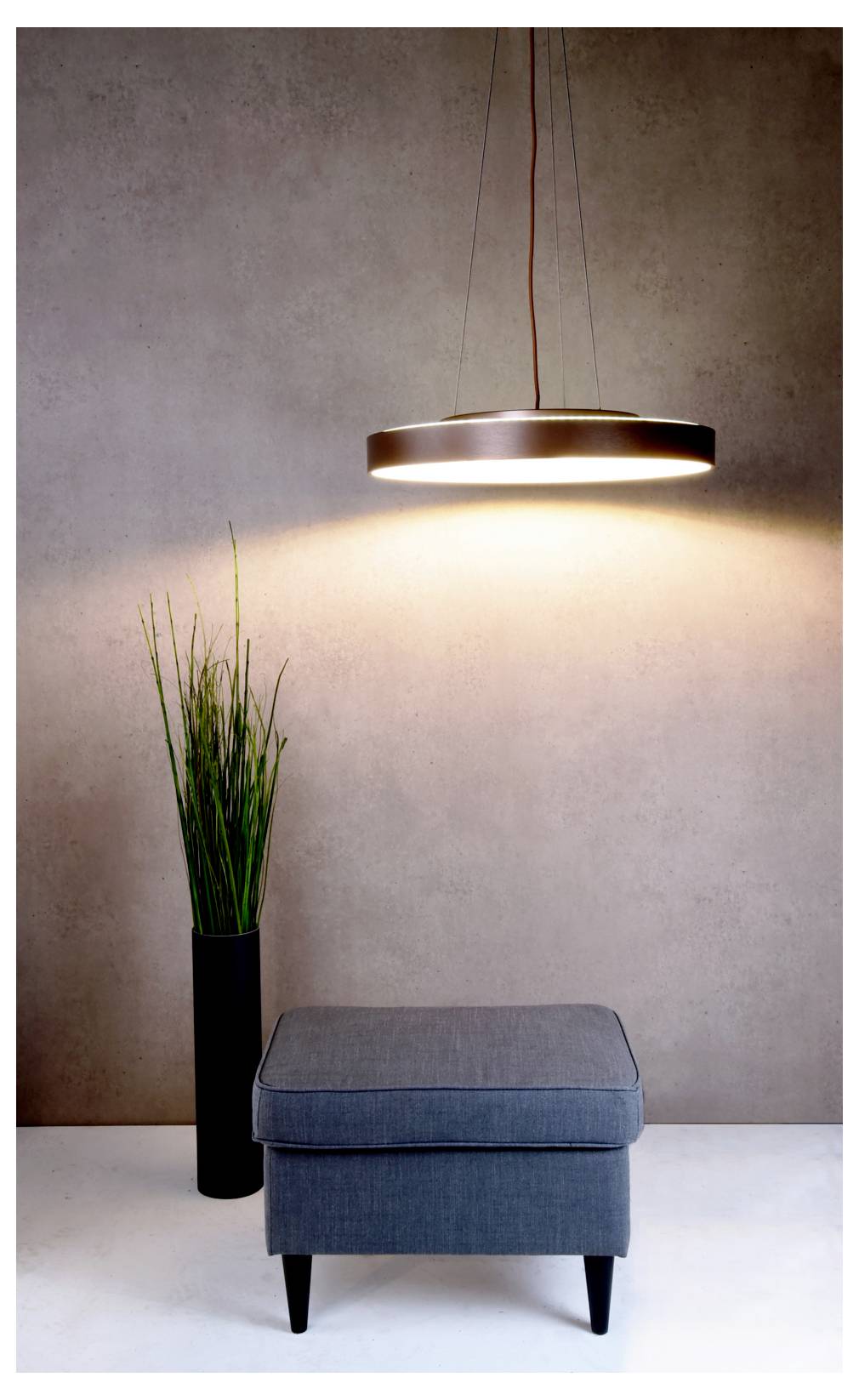 Minimal interior with a hanging round pendant light above a blue upholstered ottoman and a tall potted grass plant against a plain wall.