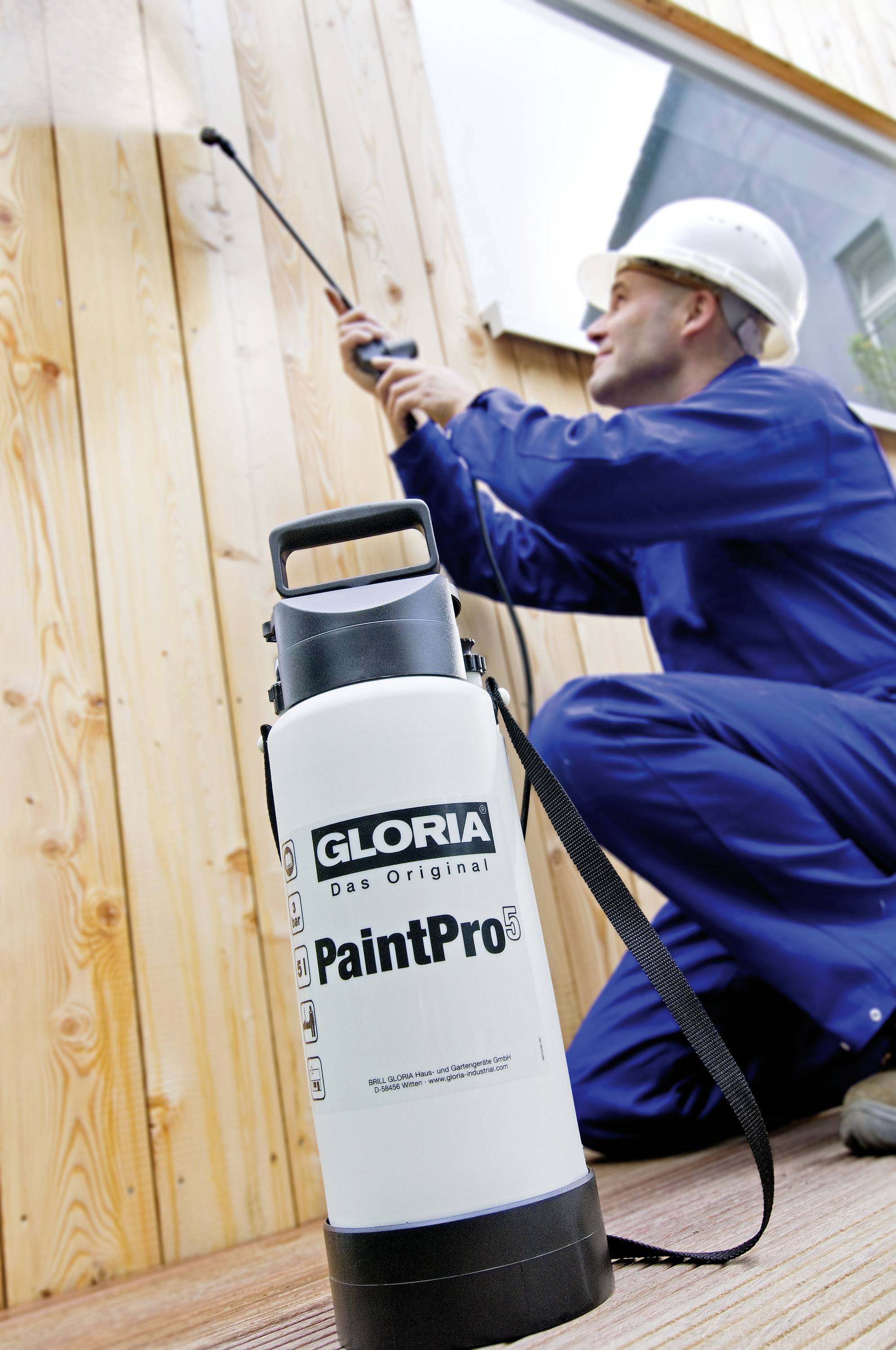 A worker in a blue uniform and hard hat is spraying wood with a device. In the foreground, a 'GLORIA PaintPro' container is standing.