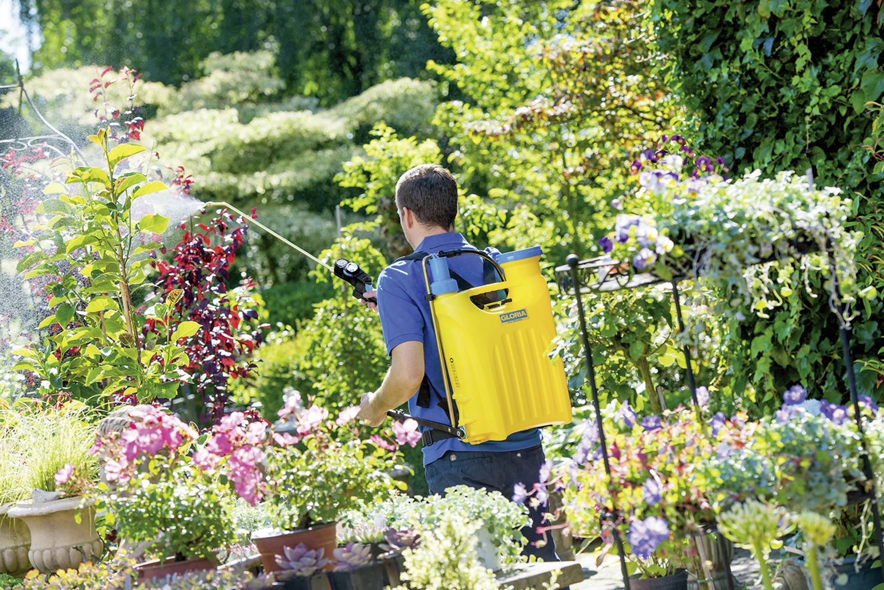 A man with a yellow backpack sprayer is watering plants in a blooming garden. Colourful flowers and green foliage surround him.