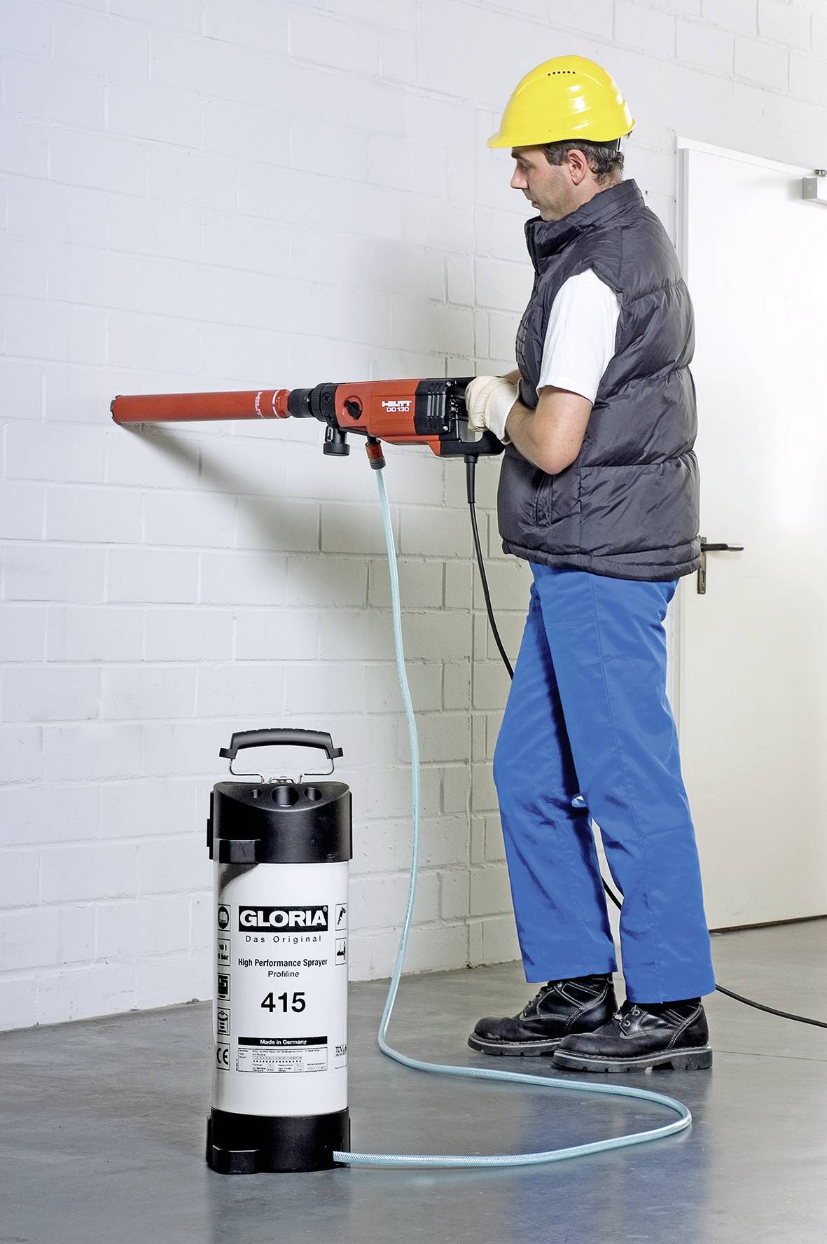 A man in protective clothing is drilling a hole in a white brick wall. A pressurised container with a hose is standing next to him on the floor.