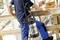A worker on a construction site carries a metal tank with a hose on their back while touching a wooden handrail, with scaffolding in the background.