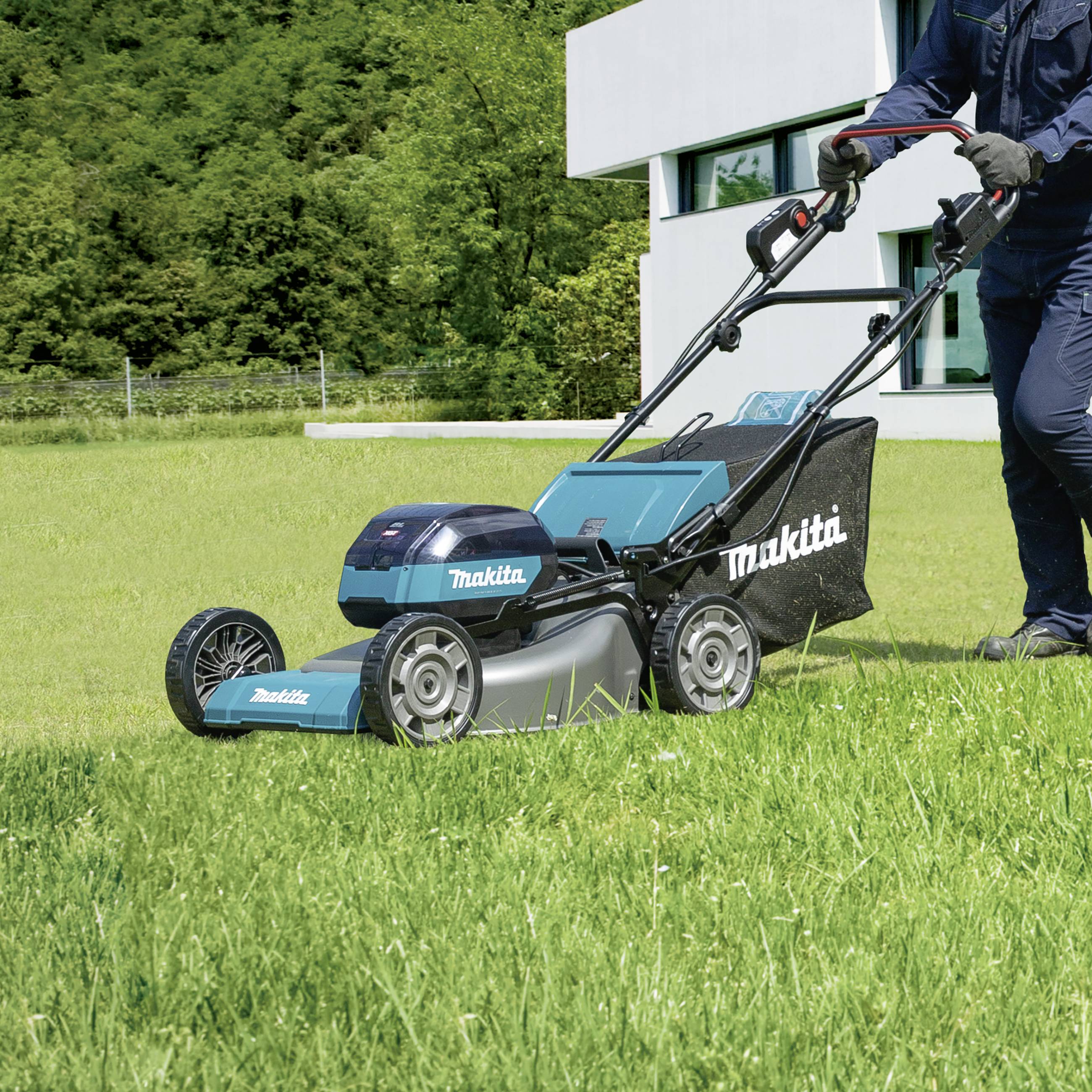 A person is mowing the lawn with a blue Makita lawnmower next to a modern white building.