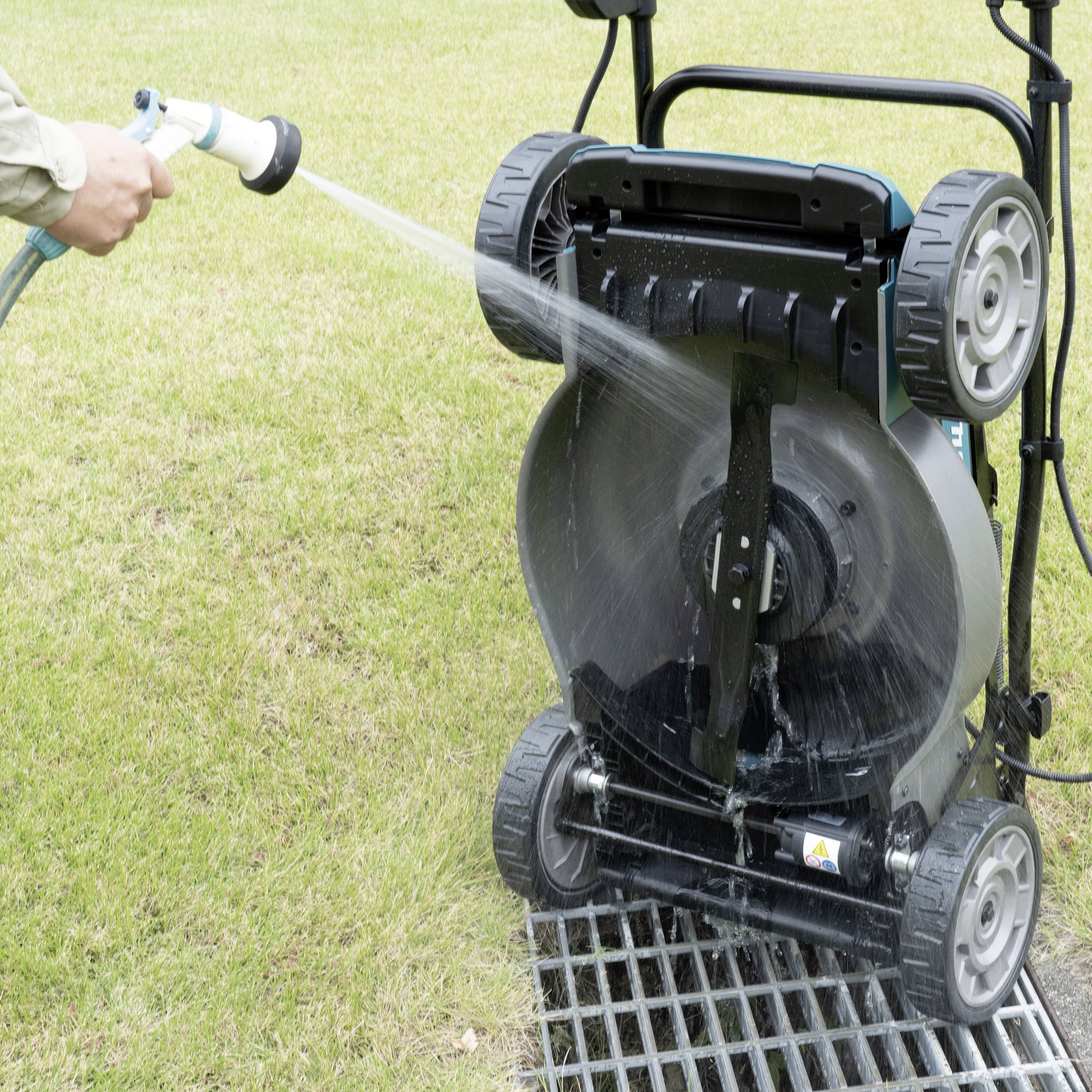 A person is cleaning the lawnmower from underneath with a hosepipe on a lawn.