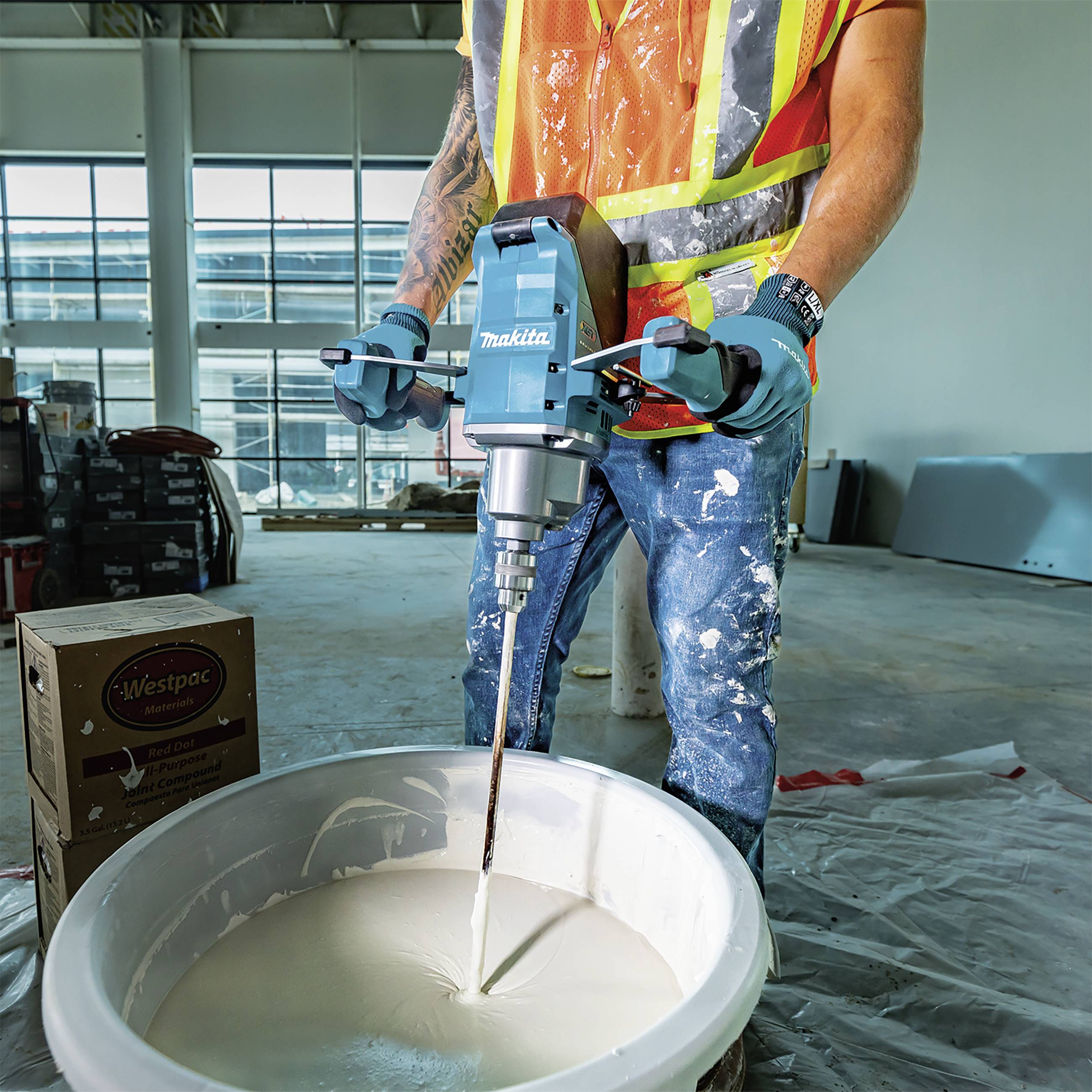 A construction worker in workwear is mixing cement with an electric mixing tool in a large bucket on a building site.