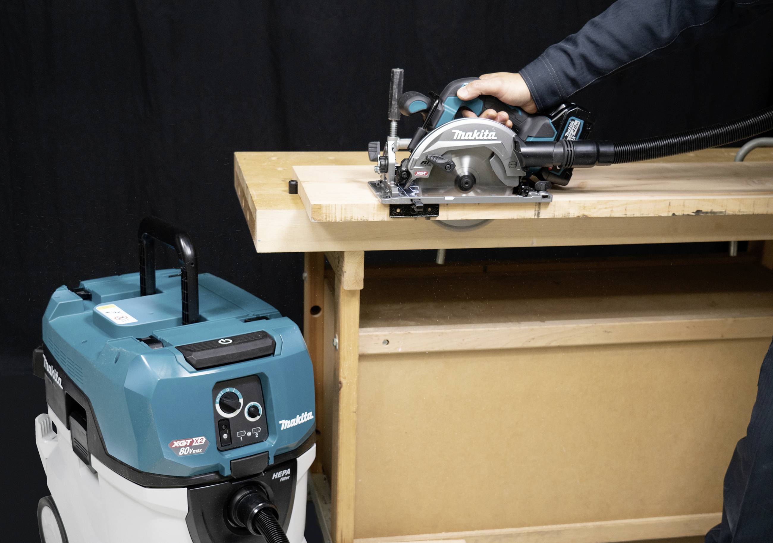 A person is using a Makita circular saw on a wooden table. A vacuum cleaner is standing ready beside the table.