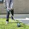 A person is using a blue lawn trimmer to cut grass next to a brick building. They are wearing blue jeans and gloves.