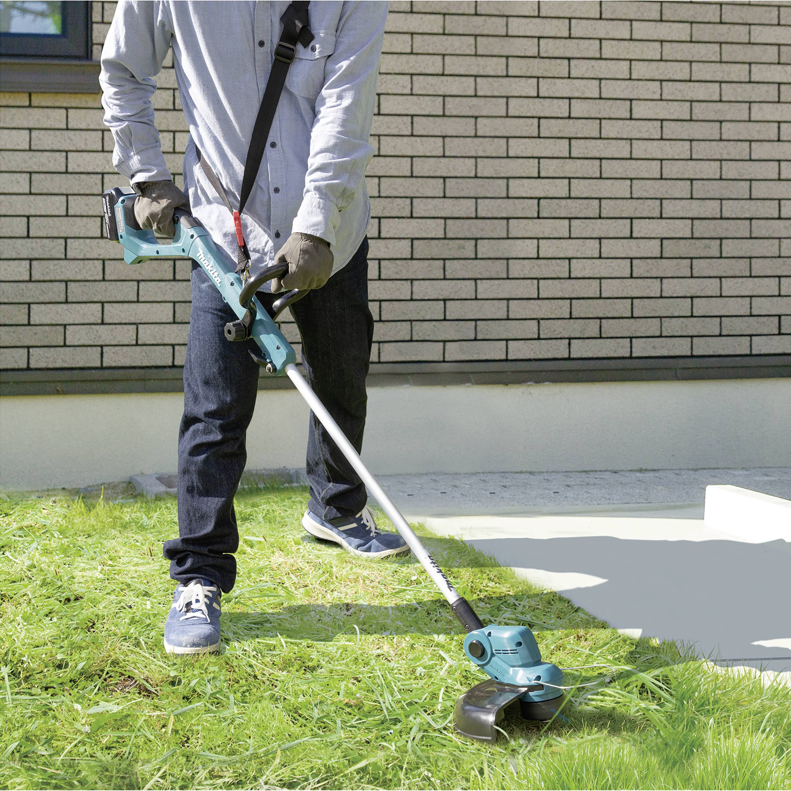 A person is using an electric lawn trimmer to cut grass near a stone wall. They are wearing gloves and jeans.