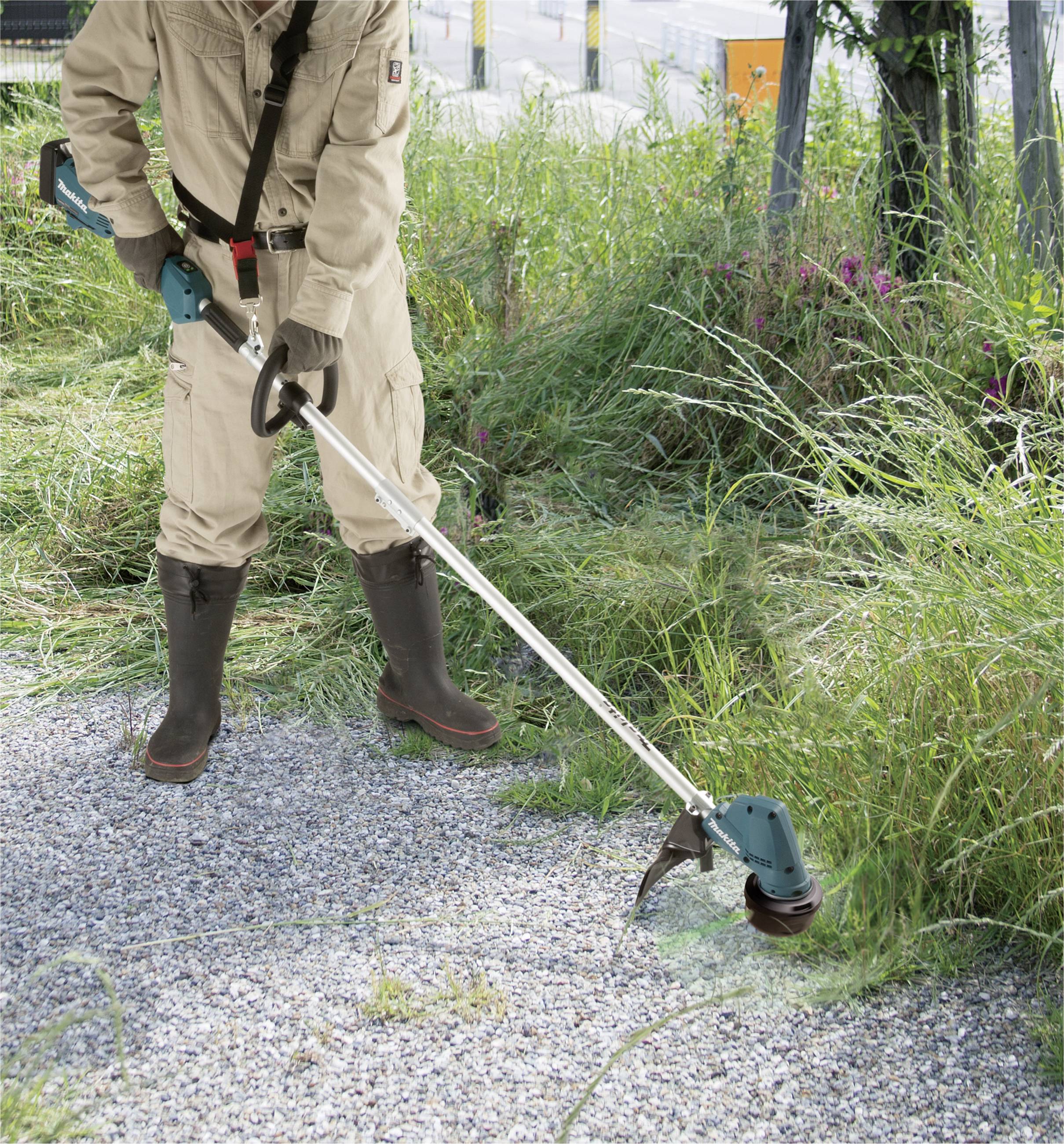 A person is standing outdoors and cutting tall grass along a gravel path using an electric strimmer.