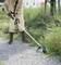 A person is standing outdoors and cutting tall grass along a gravel path using an electric strimmer.