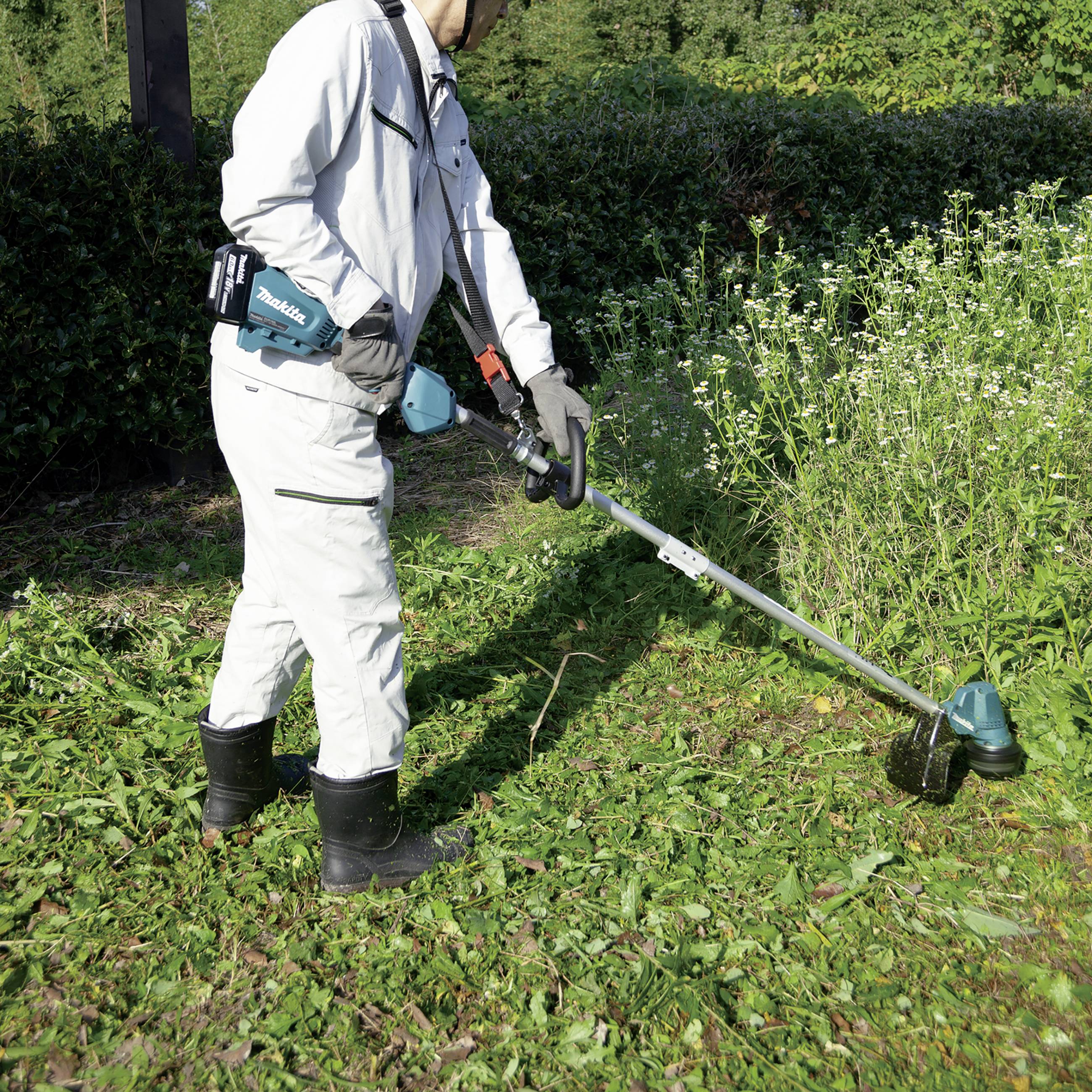 A person in protective clothing is using a strimmer to cut grass and weeds in a garden. Bushes and meadows can be seen in the background.