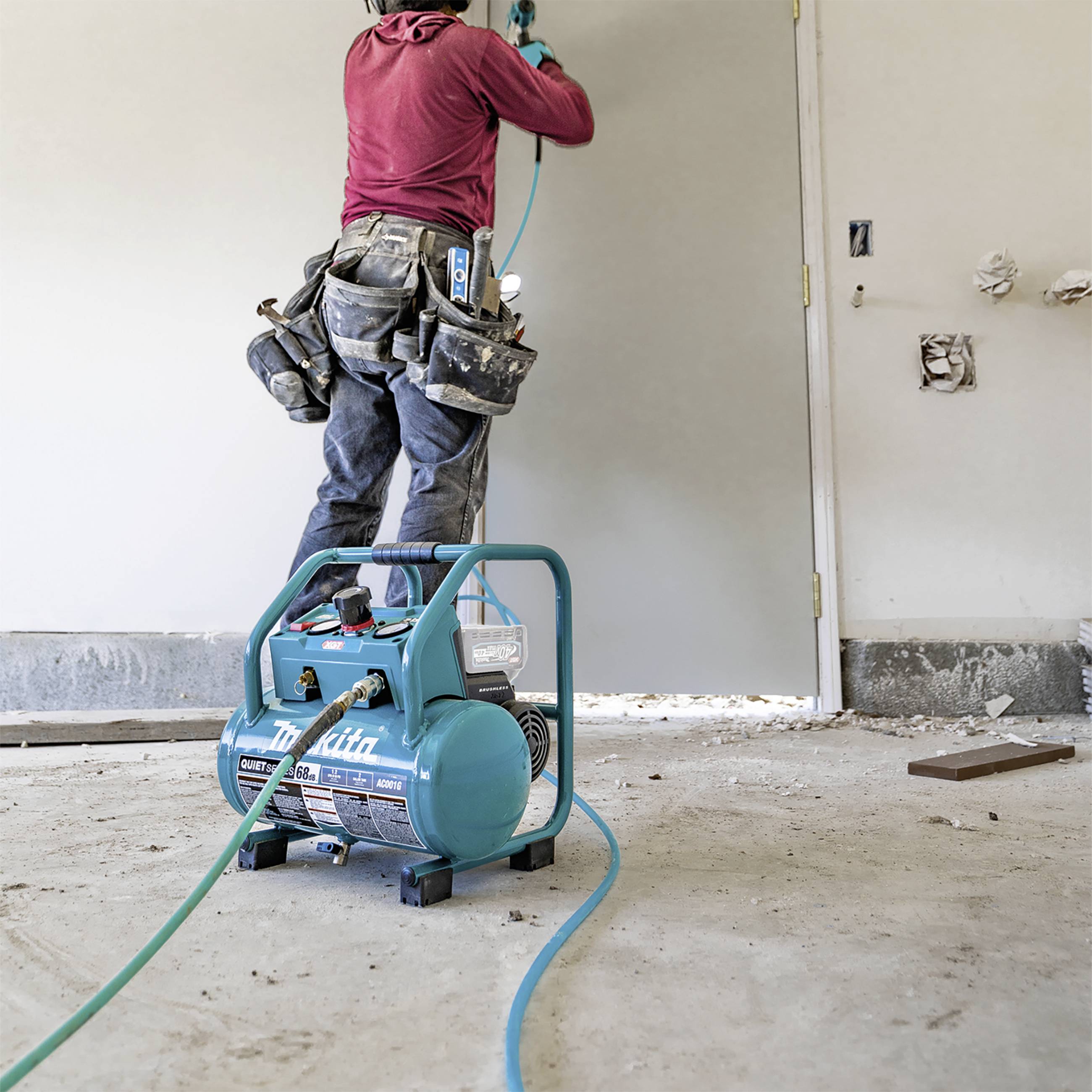 A construction worker in dirty workwear is using an air compressor to paint a door. The room is partially unfinished with exposed pipework.
