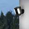 Wall-mounted security camera, switched on with bright light. Background shows blurred trees.