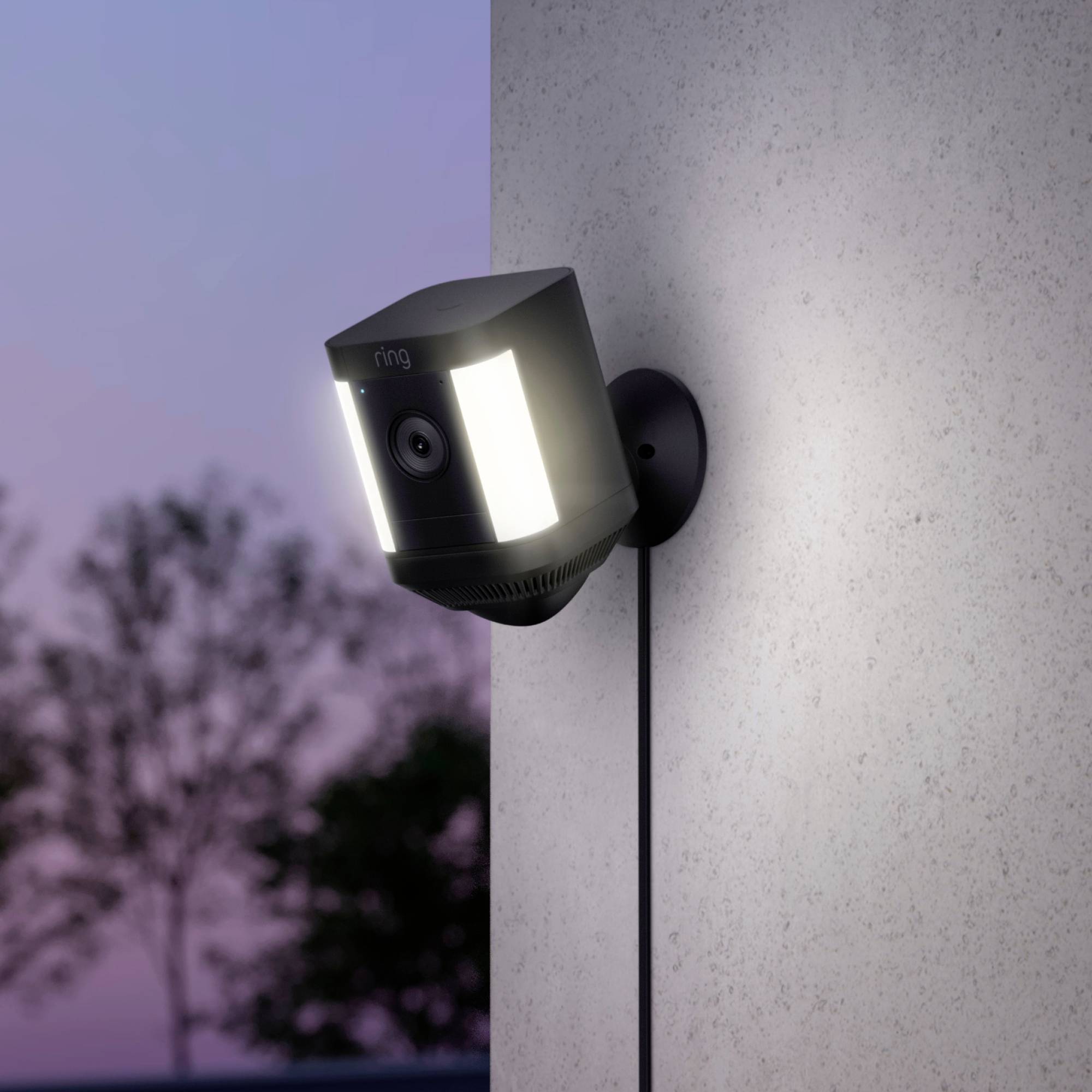 A security camera is mounted on an exterior wall. It illuminates in the twilight. Trees are visible in the background.