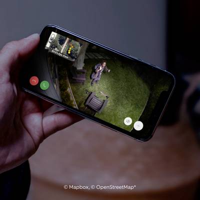 A smartphone displays an aerial view of a man in a garden via a video call app. The garden features green lawns and garden furniture.