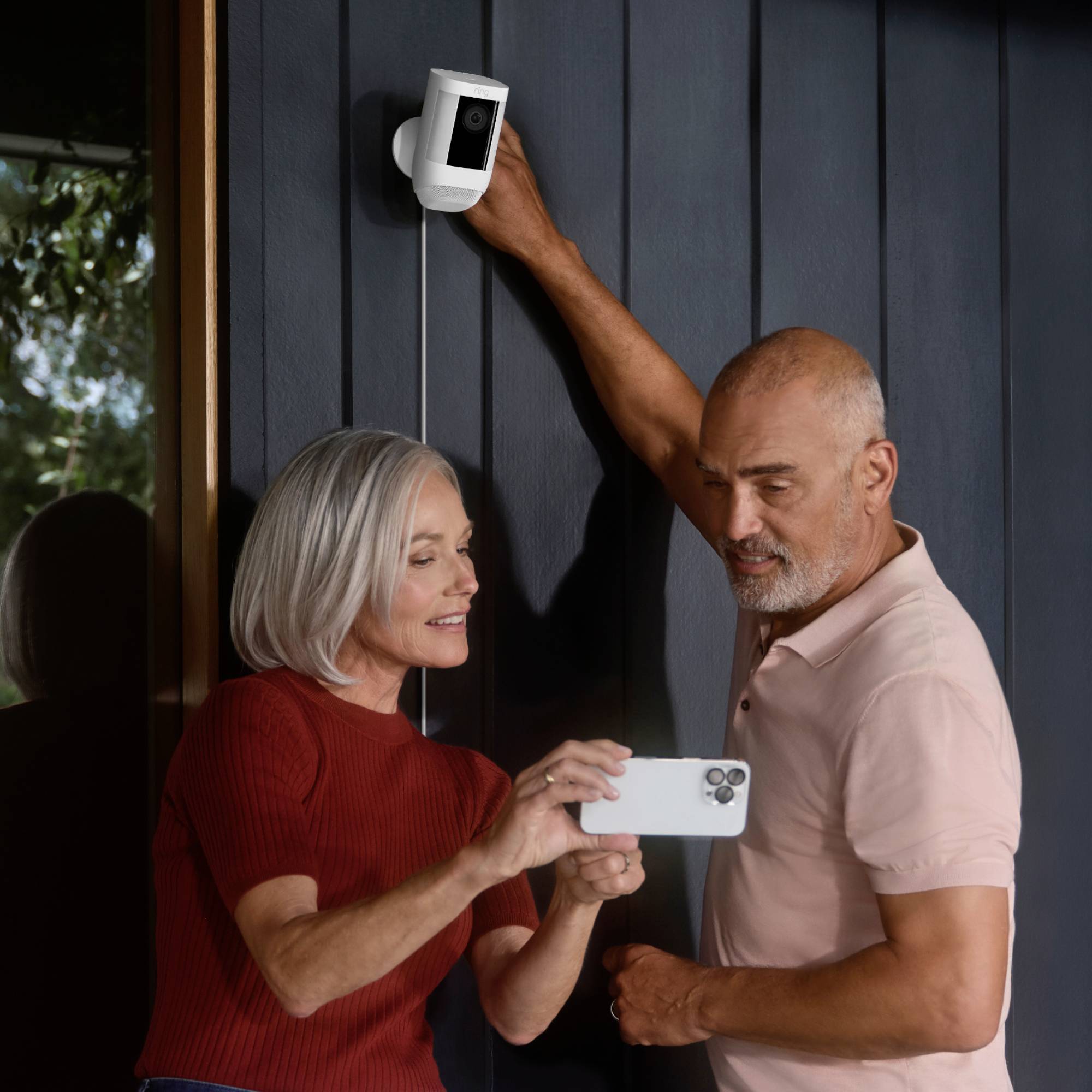 An elderly couple are installing a security camera on an external wall. The woman is holding the camera, the man is standing beside her, and both are smiling.