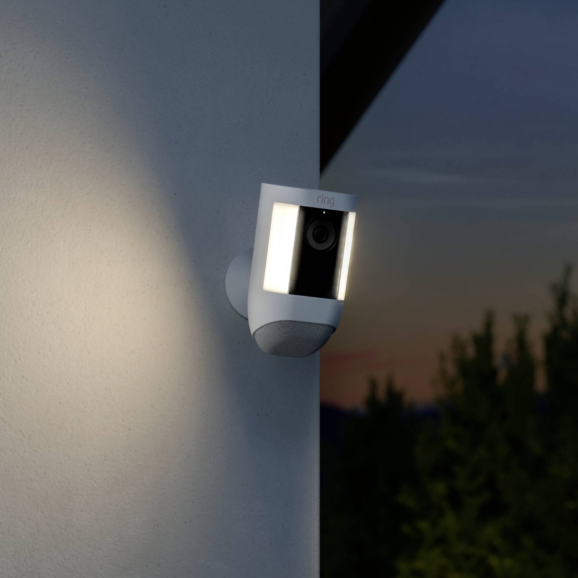 A security camera with its light on at a building during twilight. Background shows trees and a dark sky.