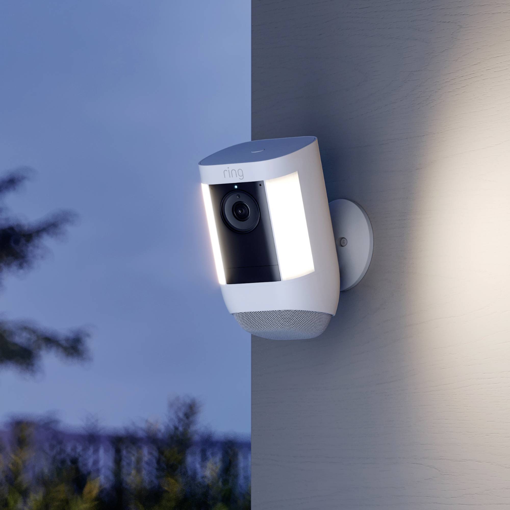 A security camera is mounted on a wall and illuminates in the twilight. Trees and the sky are visible in the background.