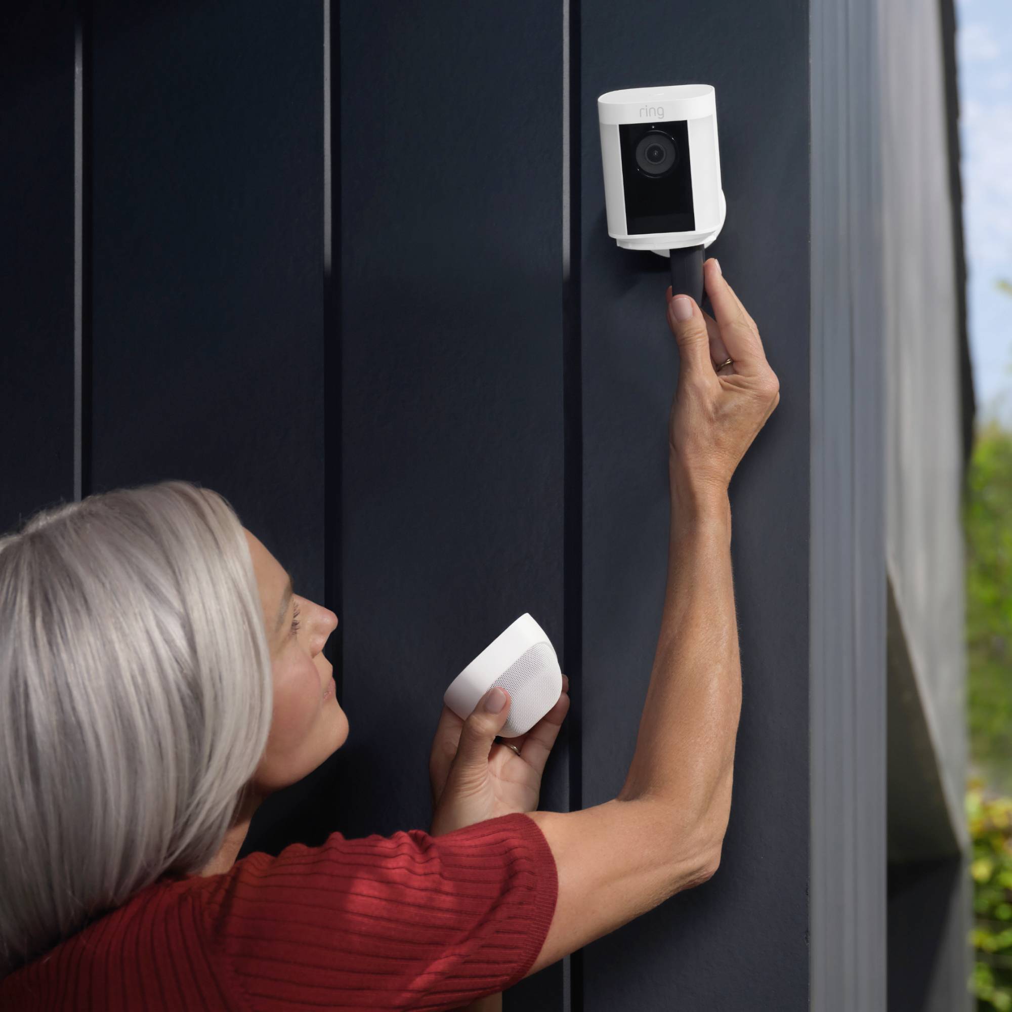 A person is installing an external surveillance camera on a dark house wall during daylight.