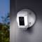 Security camera with LED light and speaker mounted on a wall, pointing outwards towards trees.