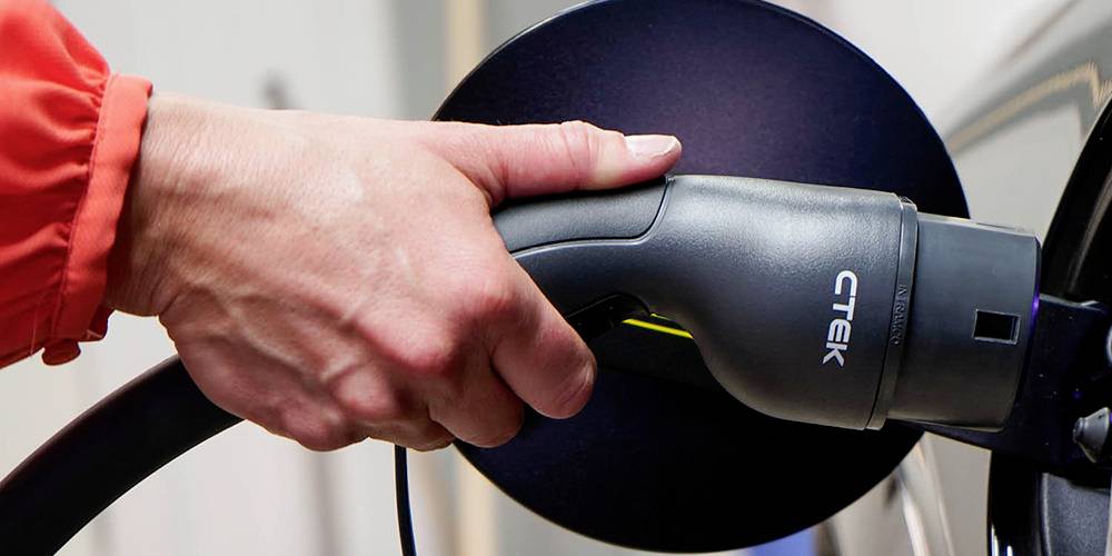 A hand is holding an electric vehicle charging plug and connecting it to a car. A print on the plug shows 'CTEK'.