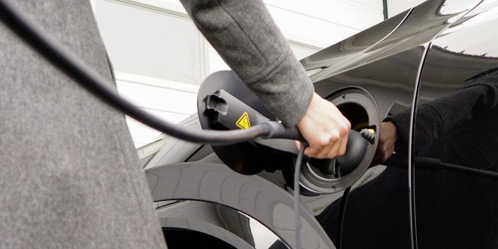 A person is charging an electric vehicle by plugging the charging cable into the car's charging socket, close-up detail of hand and cable.