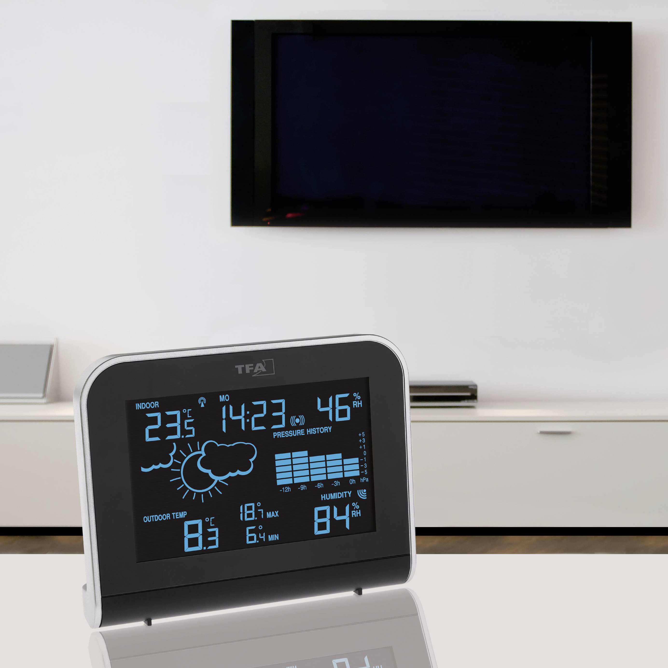 Digital weather display showing indoor and outdoor temperatures, humidity, and weather forecast in front of a television on a table.