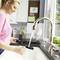 A woman in a kitchen is filling a water jug at the sink. Plants are standing on the windowsill, and crockery is drying in the background.