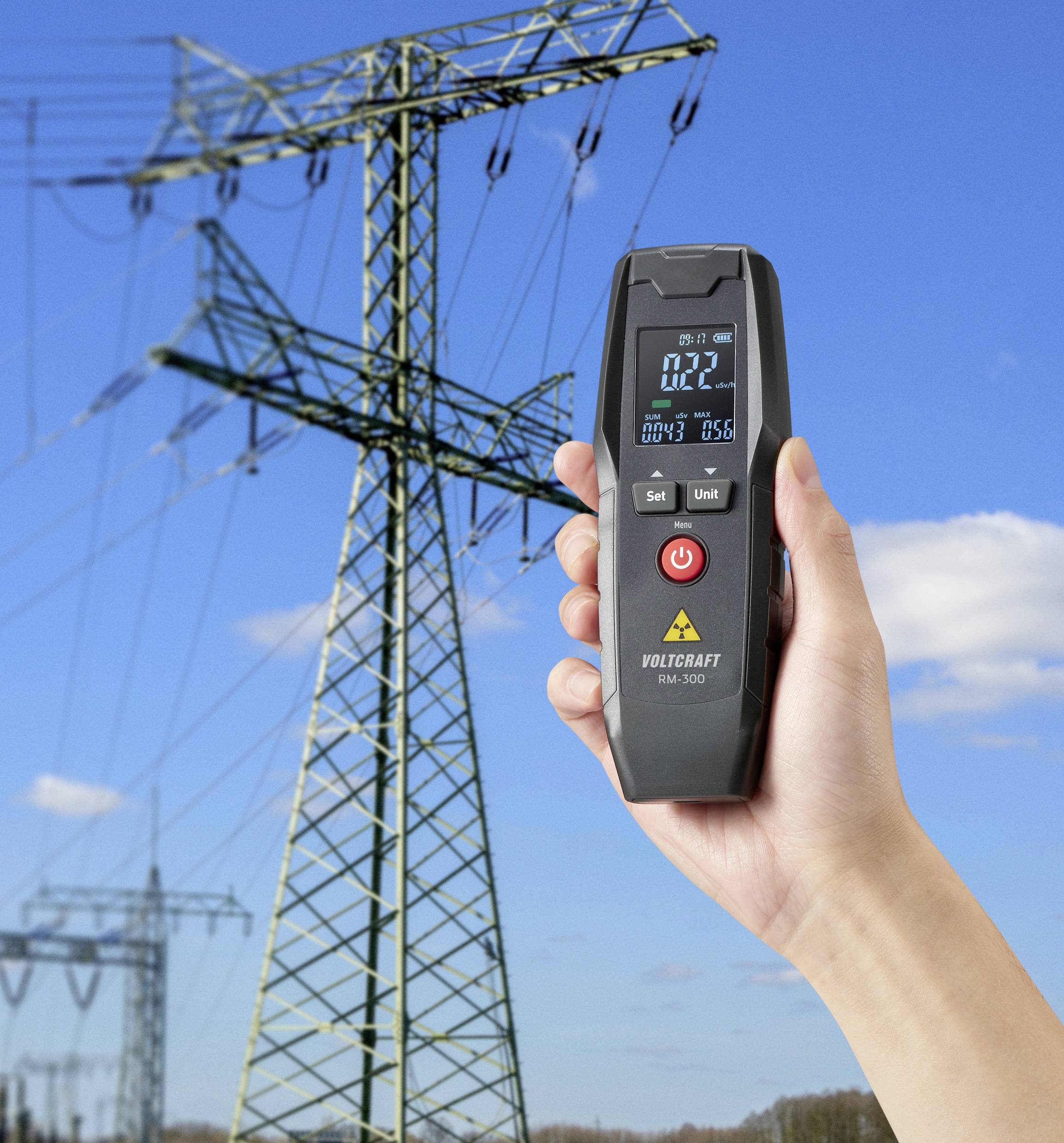 A hand holds a digital measuring device with a numerical display in front of a background featuring a large electricity pylon under a blue sky.