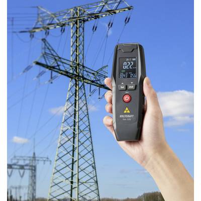 A hand holds a digital measuring device with a numerical display in front of a background featuring a large electricity pylon under a blue sky.