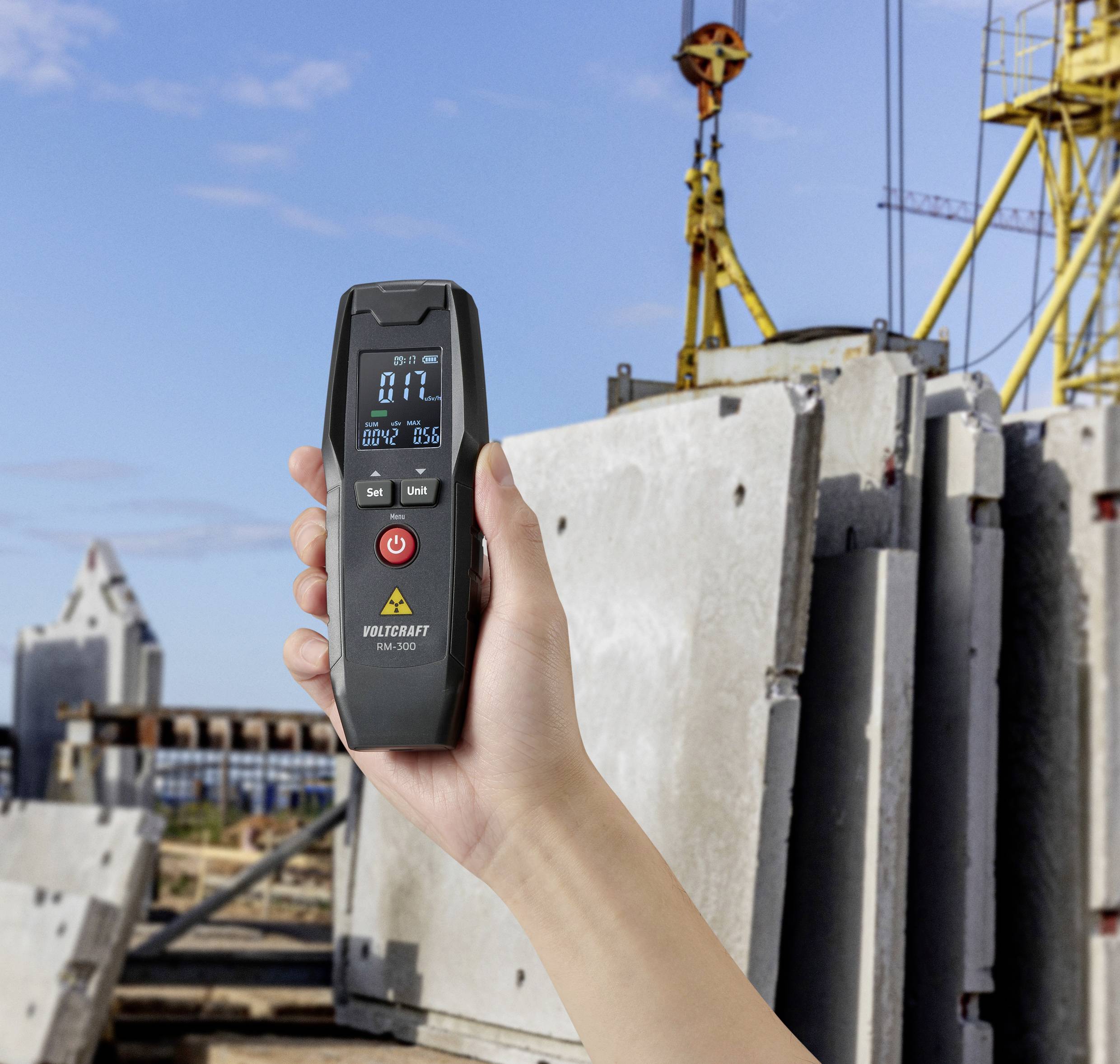 A hand holds a digital measuring device in front of a construction site with concrete slabs and cranes in the background.