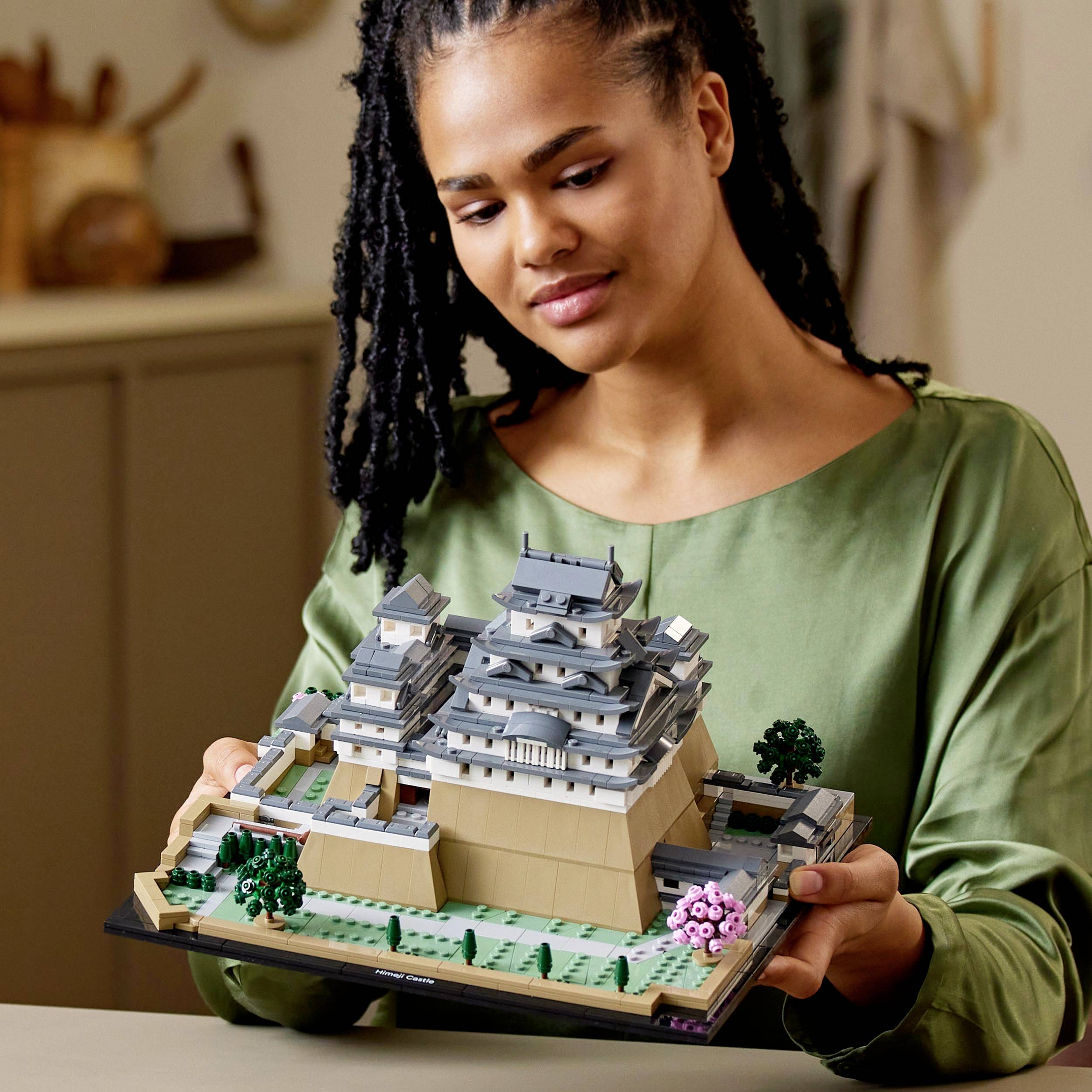 A woman is holding a Japanese castle model made of building blocks. Home accessories can be seen in the background.