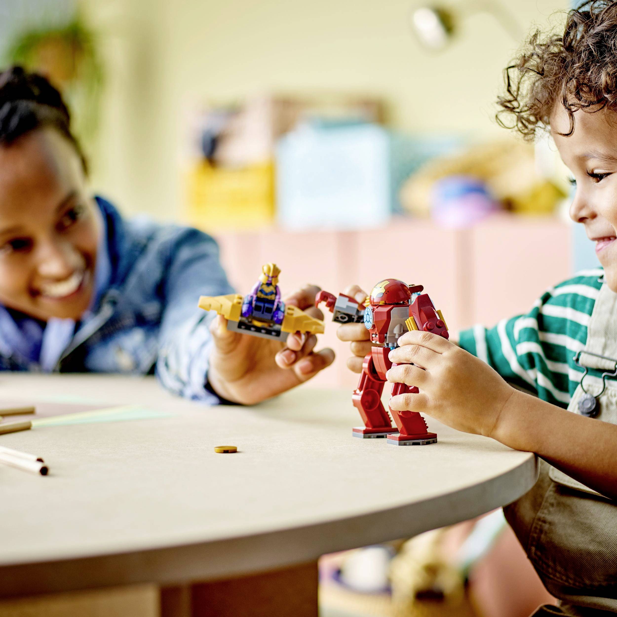 A child plays with toy robots on a table while an adult watches with a smile. Living room in the background.