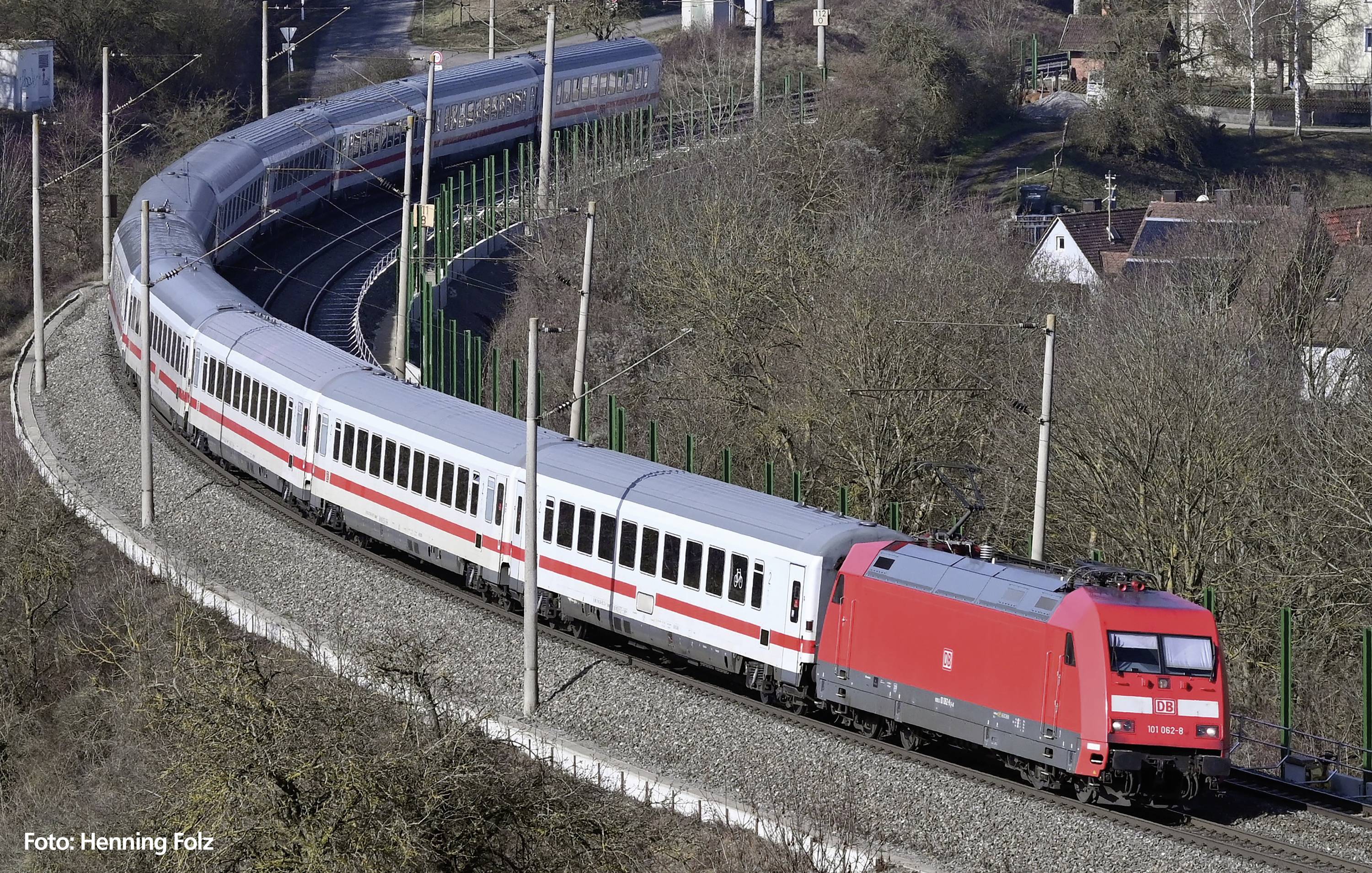A red train travels along a winding route through a rural landscape with trees and a few houses in the background.