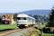 A Deutsche Bahn train travels through a rural landscape with a house in the background and hills on the horizon.