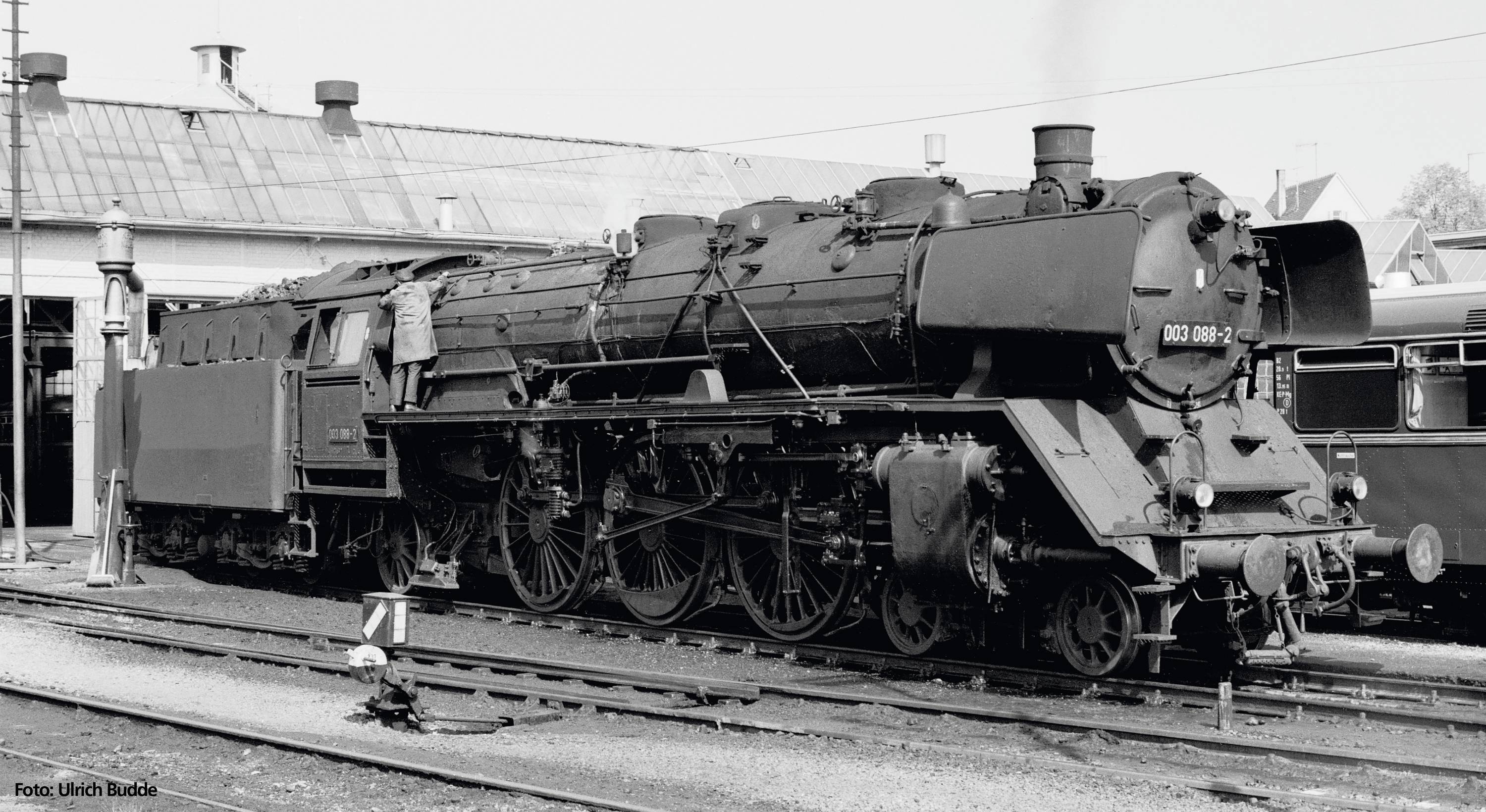 Black and white photograph of a steam locomotive on a railway track. A worker is standing beside the locomotive. A building is visible in the background.
