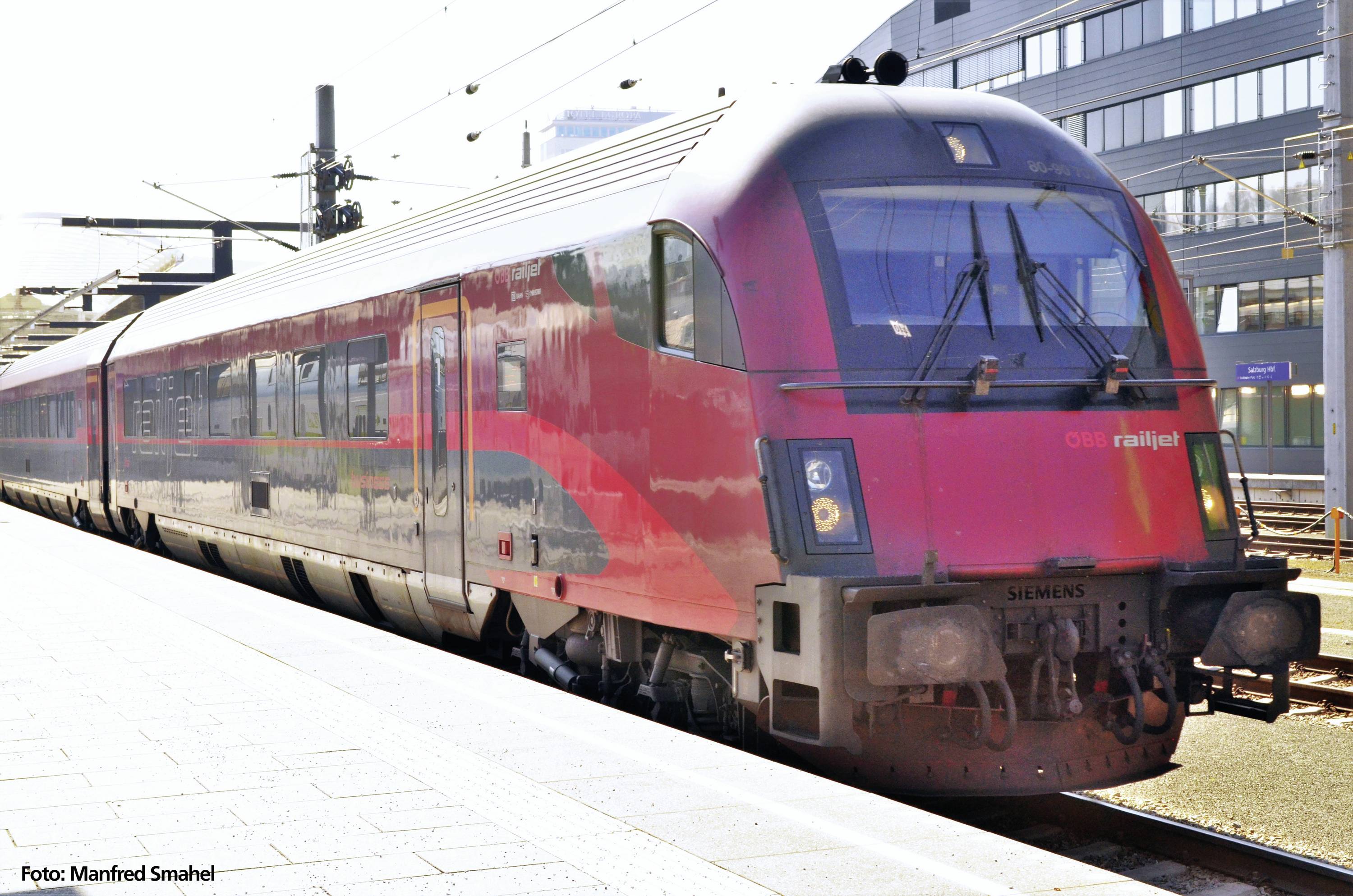 A red ÖBB Railjet train is standing at a platform in sunny weather. It is modern in design and ready for departure.