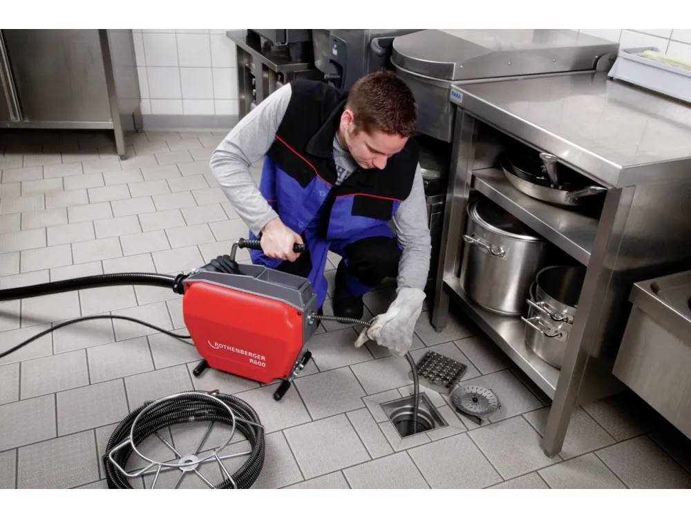A person is using a plumbing tool to clean a drain in a commercial kitchen, surrounded by stainless steel countertops and tiled flooring.